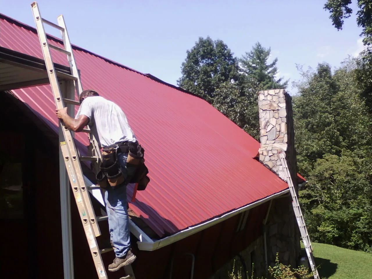 Roofer on a ladder beside a red metal roof and stone chimney — Soffit & Fascia Repair by Roofing Brothers in Columbia Cross Roads